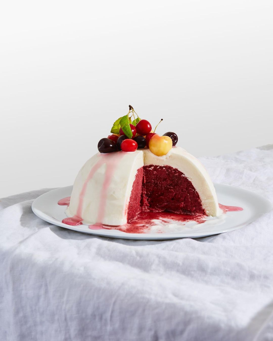 Hemispherical cake with white frosting and cherry topping, sliced to reveal a dark red filling. On white plate and textured cloth.