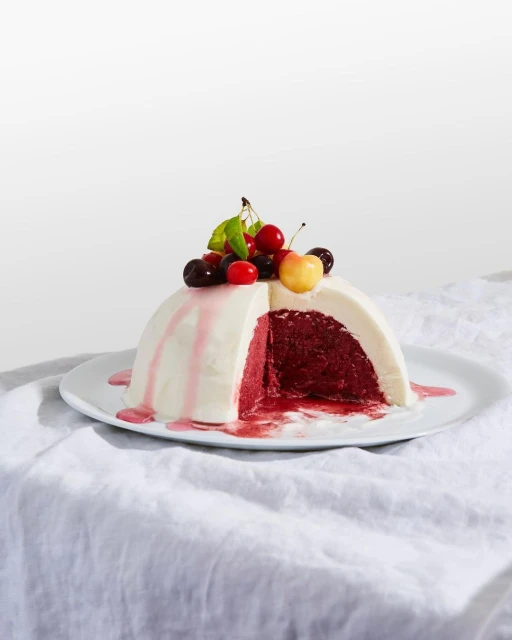 Hemispherical cake with white frosting and cherry topping, sliced to reveal a dark red filling. On white plate and textured cloth.