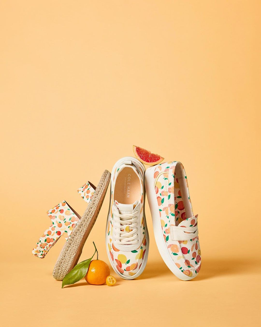 Still life of Cole Haan shoes and sandals with an orange citrus fruit, featuring a matching fruit pattern on the shoes.
