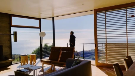 Man standing on a balcony overlooking the ocean from a modern living room with large glass windows photographed by Alpha Smoot.