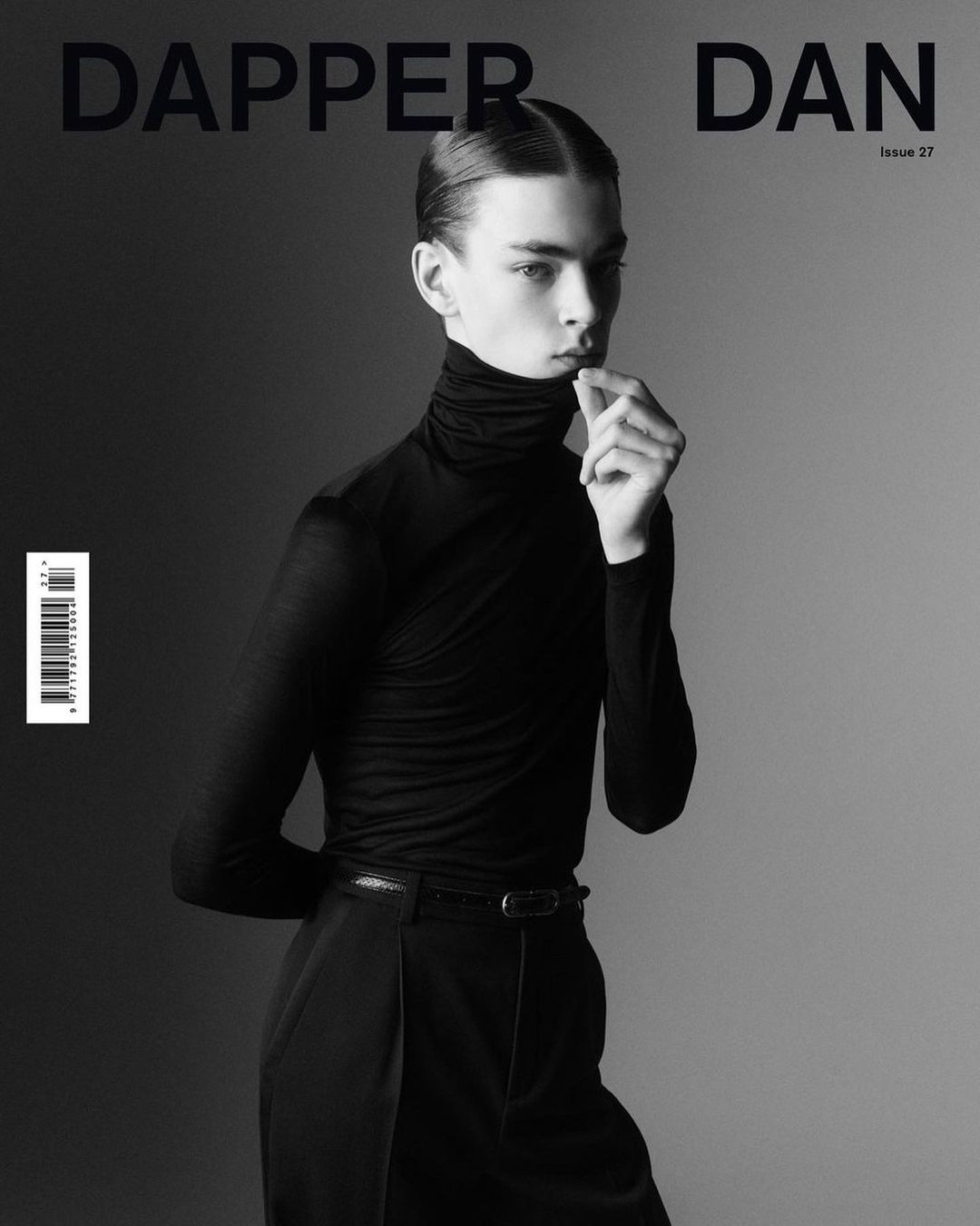 Black and white studio shot of a male fashion model wearing a black turtleneck and trousers on a gradient grey background.