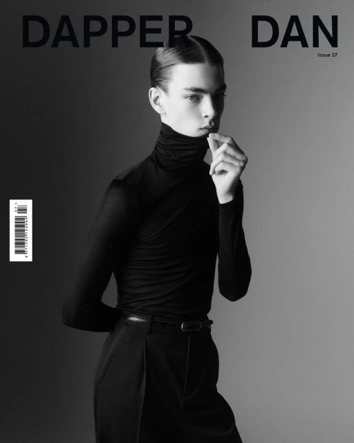 Black and white studio shot of a male fashion model wearing a black turtleneck and trousers on a gradient grey background.