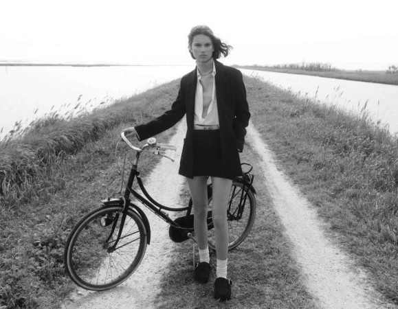A woman in a black blazer and shorts stands with a bicycle on a gravel path between grassy banks, captured in monochrome.