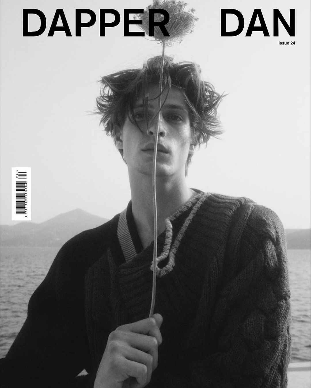 Black and white portrait of a young man holding a tall thin flower in front of his face, wearing a dark knit sweater outdoors by water.