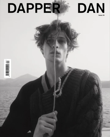 Black and white portrait of a young man holding a tall thin flower in front of his face, wearing a dark knit sweater outdoors by water.