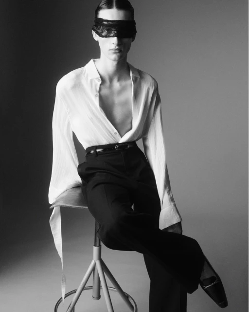 Monochrome studio portrait of an adult woman with a dark blindfold, wearing a white unbuttoned shirt and dark pants, sitting on a stool.