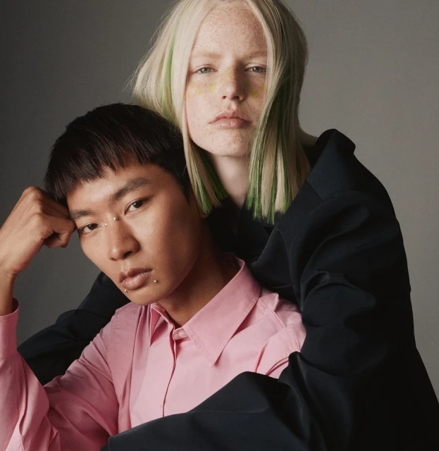 Two people in a close studio shot. One with pink shirt and another with a black jacket and blonde hair with green highlights.