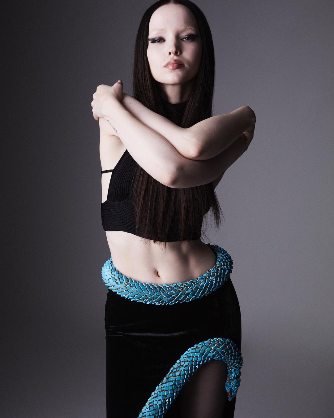 Portrait of a woman in a black top and skirt with a turquoise snake prop wrapped around her body, posing against a grey backdrop.