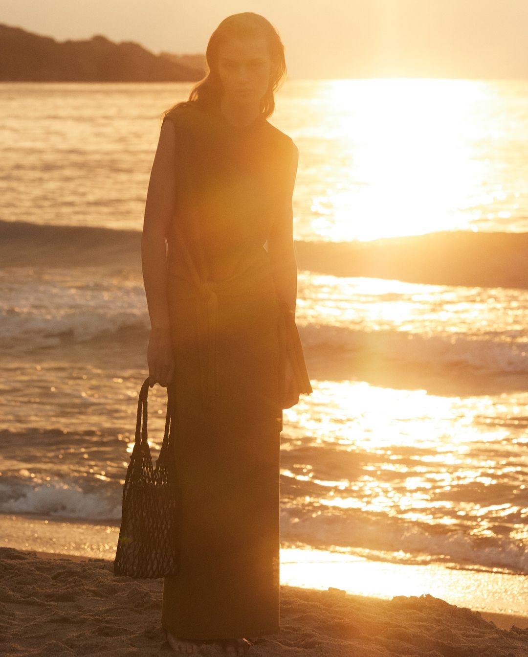 An adult woman stands on a sandy beach at sunset, wearing a sleeveless dress and holding a mesh bag, with the ocean in the background.