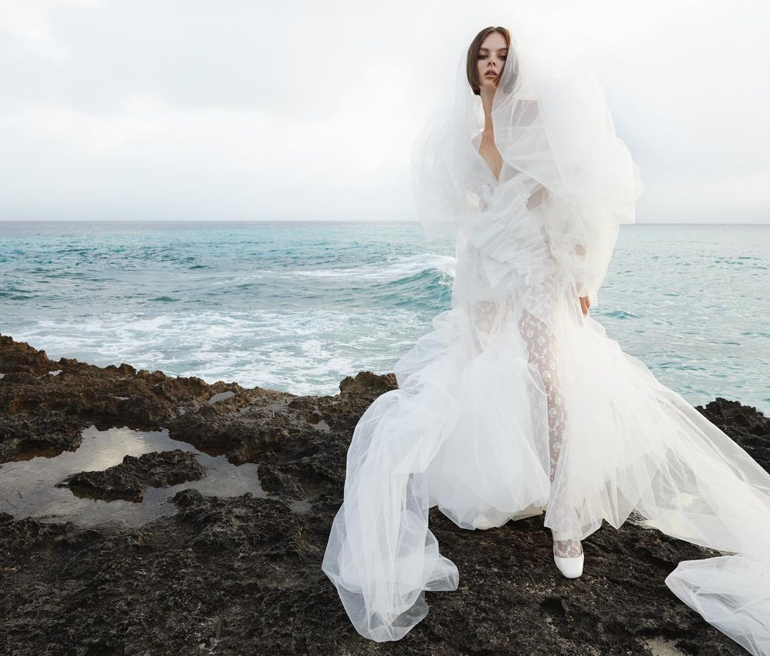 Woman poses in ruffled white dress on rocky beach. The ocean waves and cloudy sky create a dreamy backdrop. Fashion editorial.