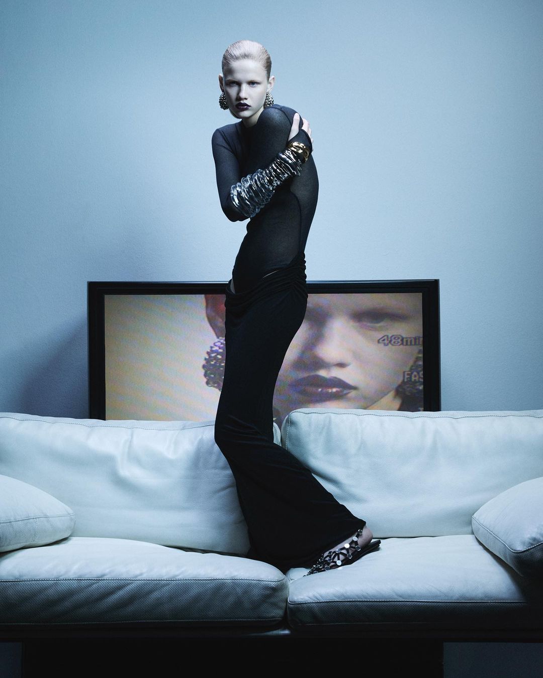 Fashionable woman in a black outfit strikes a pose on a white couch against a pale blue wall, with a television behind her.