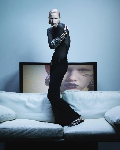 Fashionable woman in a black outfit strikes a pose on a white couch against a pale blue wall, with a television behind her.