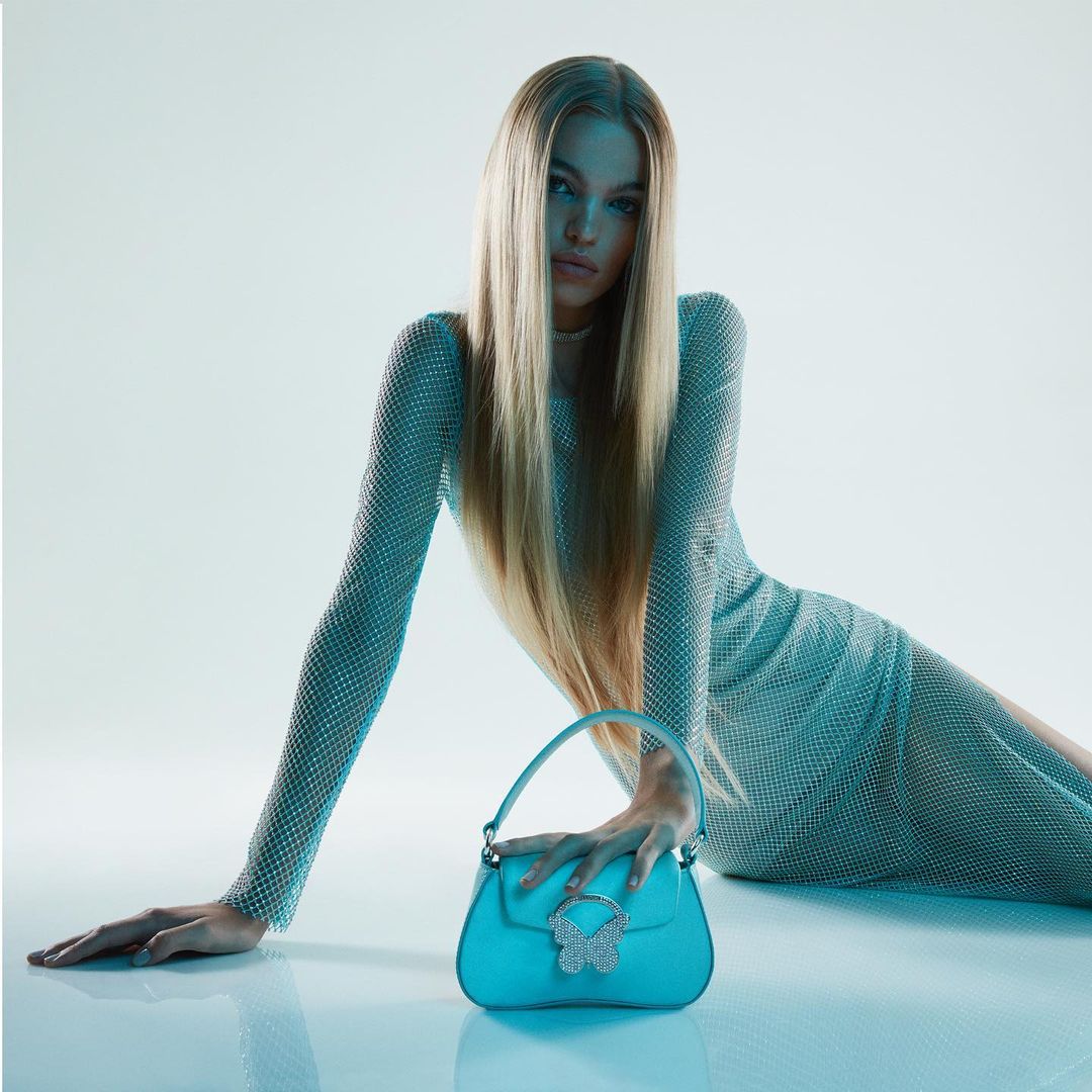 Fashion shot of a blonde woman in a glittering dress, posing with a turquoise handbag adorned with a butterfly embellishment.