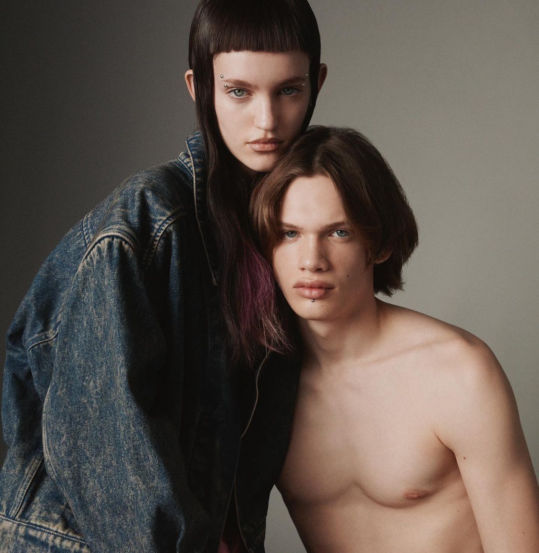 Portrait of a female model wearing an oversized denim jacket standing behind a shirtless male model against a grey studio background