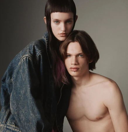 Portrait of a female model wearing an oversized denim jacket standing behind a shirtless male model against a grey studio background