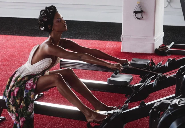 Glamorous adult female works out on a rowing machine in a gym setting, wearing an evening gown and hair rollers for a surreal effect.