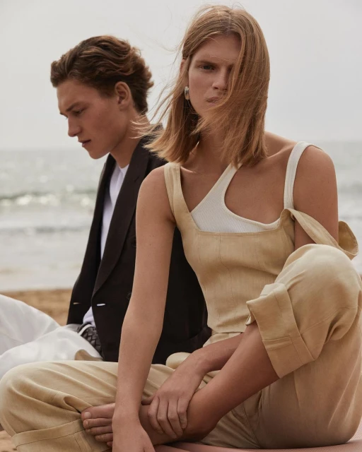 Two fashion models, an adult male and an adult female, sit on a sandy beach with the ocean in the background. Editorial fashion shot.