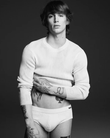 Monochrome portrait of a tattooed man wearing a white knit sweater and briefs against a dark background
