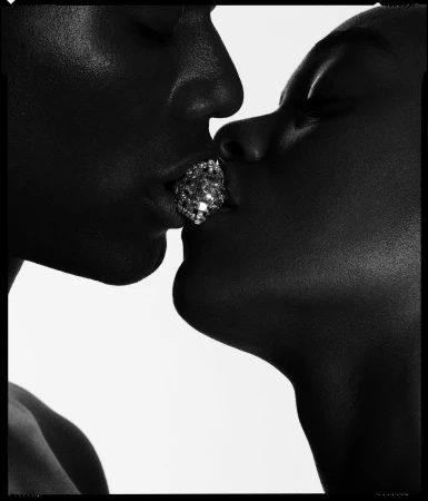 Close-up black and white image of two dark-skinned adults kissing, with a large, multifaceted, diamond-like object visible at point of contact.