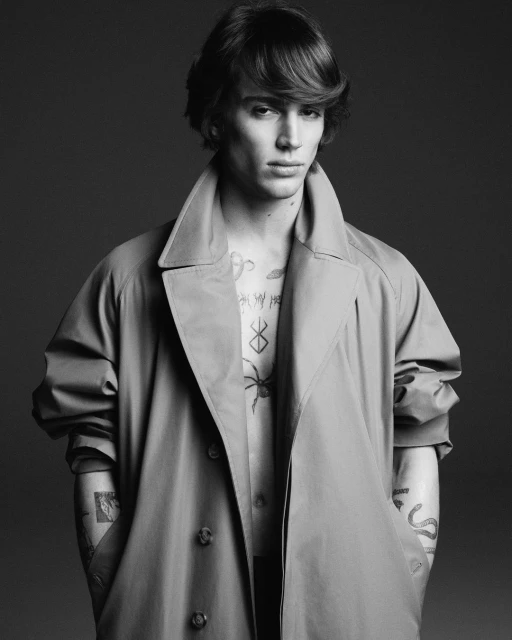 Black and white portrait of a young adult male with medium length brown hair, wearing an open trench coat with tattoos on his chest and arms.
