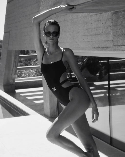 Fashion shot of a model wearing a black swimsuit and sunglasses, posing against a white concrete wall in a monochrome outdoor setting.
