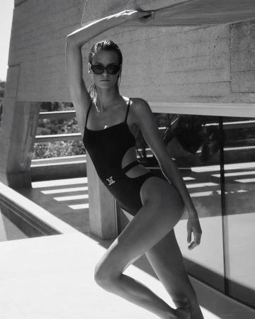Fashion shot of a model wearing a black swimsuit and sunglasses, posing against a white concrete wall in a monochrome outdoor setting.