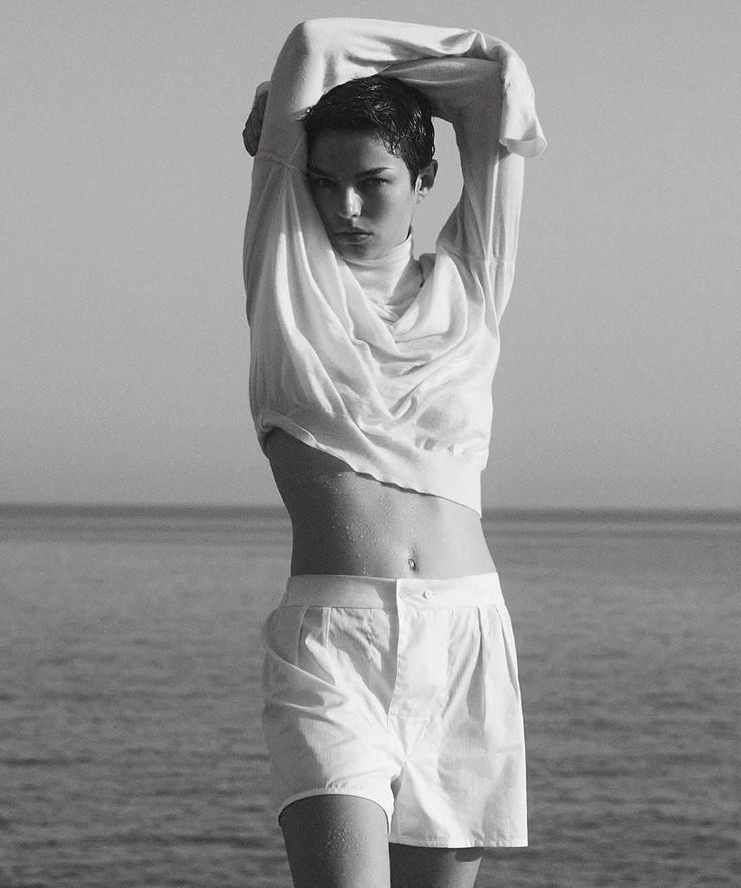 Monochrome portrait of a young woman in a white sweater and shorts standing outdoors, with a sea and sky background, captured in natural light.