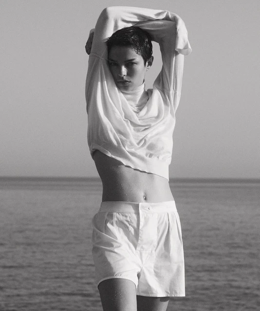 Monochrome portrait of a young woman in a white sweater and shorts standing outdoors, with a sea and sky background, captured in natural light.
