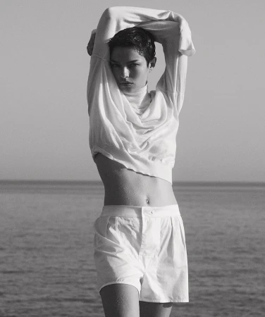 Monochrome portrait of a young woman in a white sweater and shorts standing outdoors, with a sea and sky background, captured in natural light.