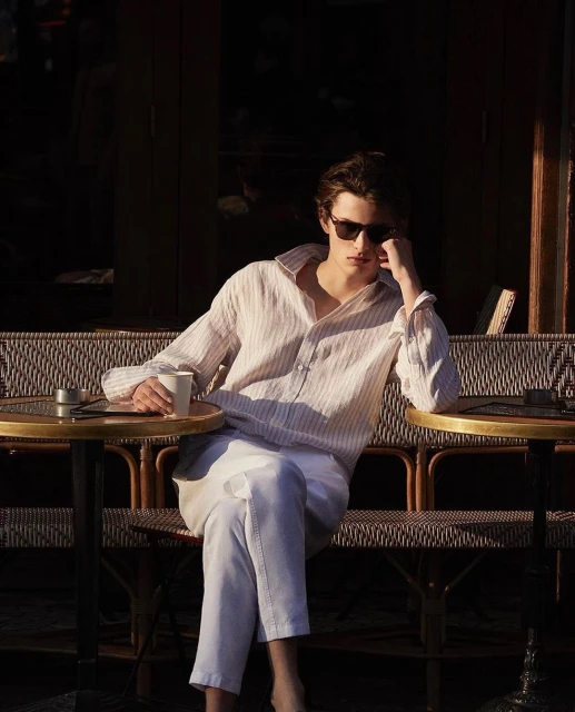 Young adult male relaxes at outdoor cafe with sunglasses, wearing unbuttoned long-sleeved striped shirt and white pants, enjoying the sunlight.