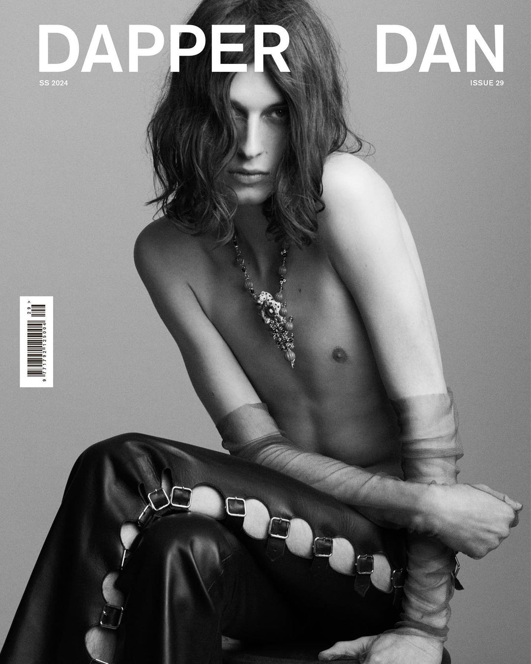 Black and white fashion shot of a young adult male with long hair wearing jewelry and leather pants, posing in front of a grey backdrop.