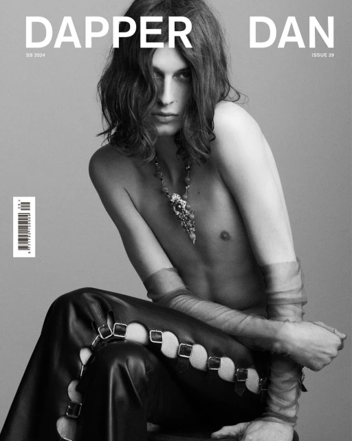 Black and white fashion shot of a young adult male with long hair wearing jewelry and leather pants, posing in front of a grey backdrop.