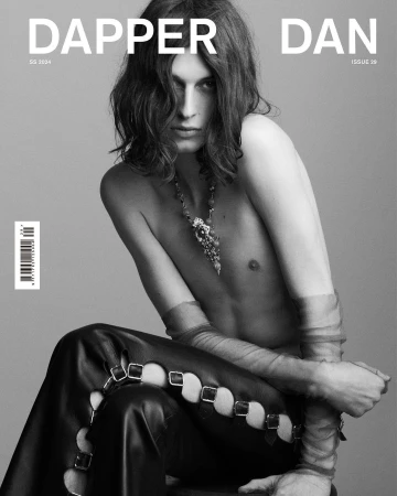 Black and white fashion shot of a young adult male with long hair wearing jewelry and leather pants, posing in front of a grey backdrop.