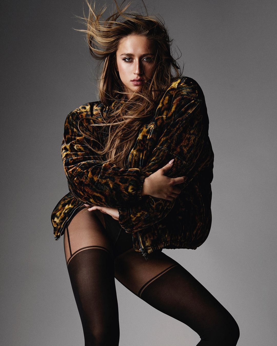 Portrait of a young adult woman with flowing brown hair, wearing a leopard print jacket and black stockings in a photo studio.