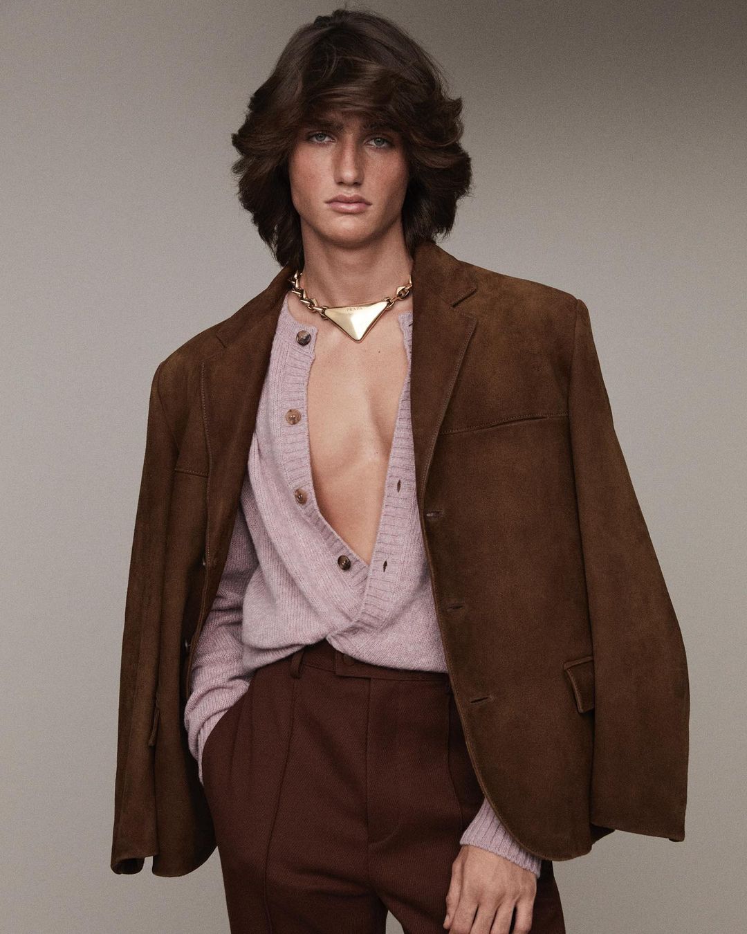 Male model with brown hair poses indoors wearing brown jacket, light pink cardigan, gold necklace and brown pants in studio lighting.