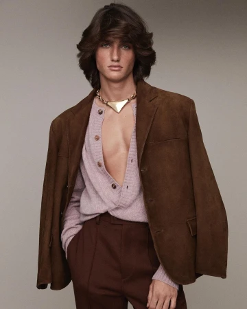 Male model with brown hair poses indoors wearing brown jacket, light pink cardigan, gold necklace and brown pants in studio lighting.
