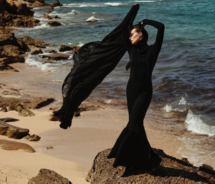 Woman in black dress and black shawl poses on a rocky beach near the ocean with waves. The shot is an eye-level, medium shot.