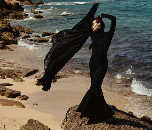 Woman in black dress and black shawl poses on a rocky beach near the ocean with waves. The shot is an eye-level, medium shot.