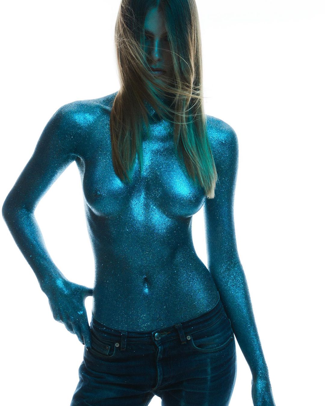 Woman covered in glitter, posing with a hand on her jeans, white background, portrait orientation, studio lighting, fashion, beauty.