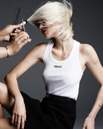 A woman in a white tank top getting her hair cut by a stylist holding scissors, set against a grey background.