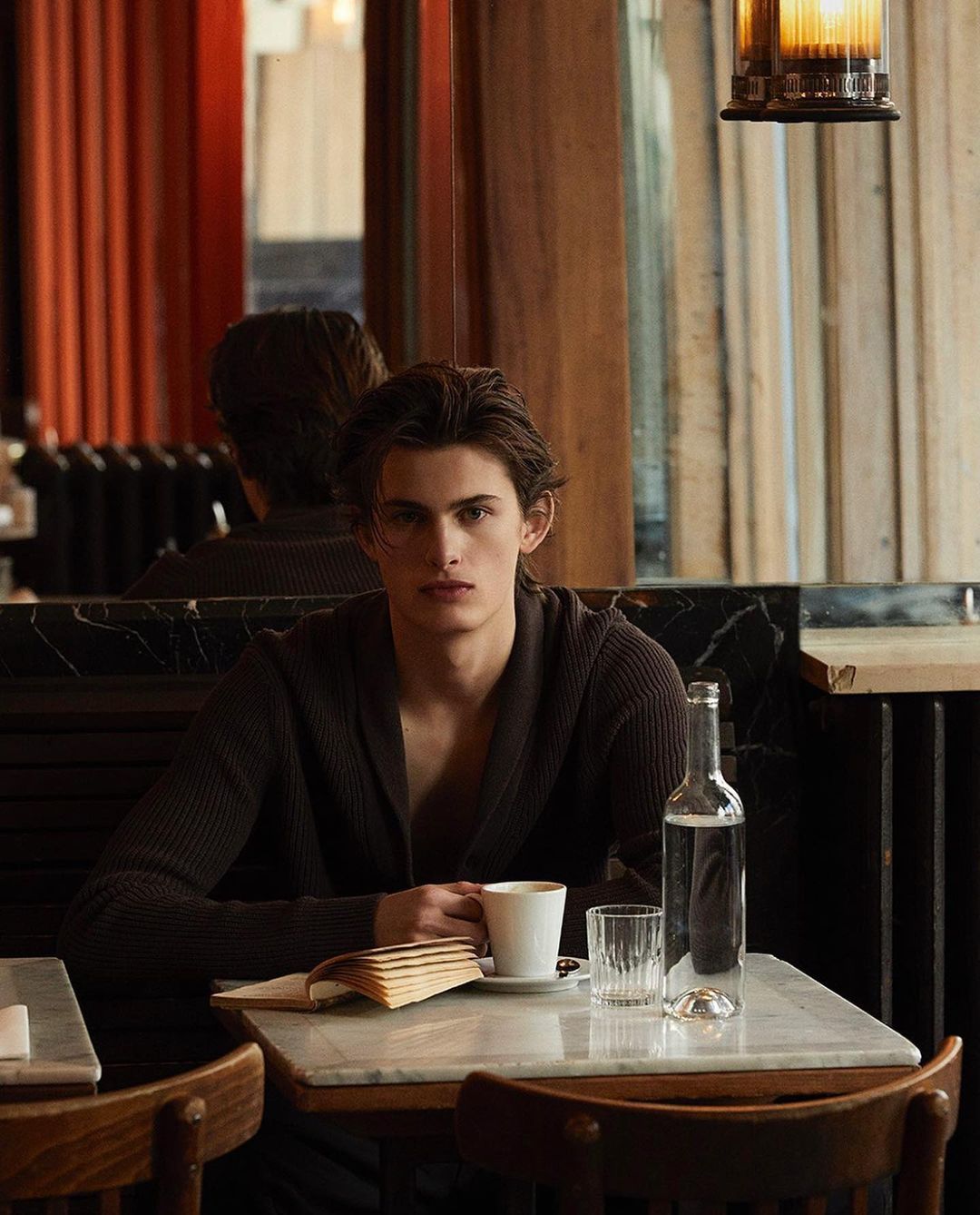 Adult male sits at a table with a book, coffee, and water in an indoor setting with muted lighting and dark brown tones.