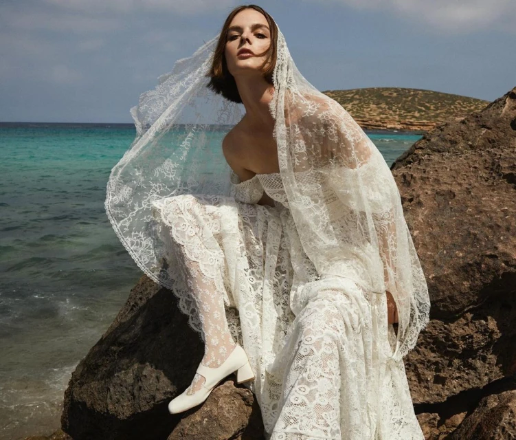 A woman in a white lace dress and veil sits on rocks by the ocean with blue water and sky in the background, creating an editorial fashion image.