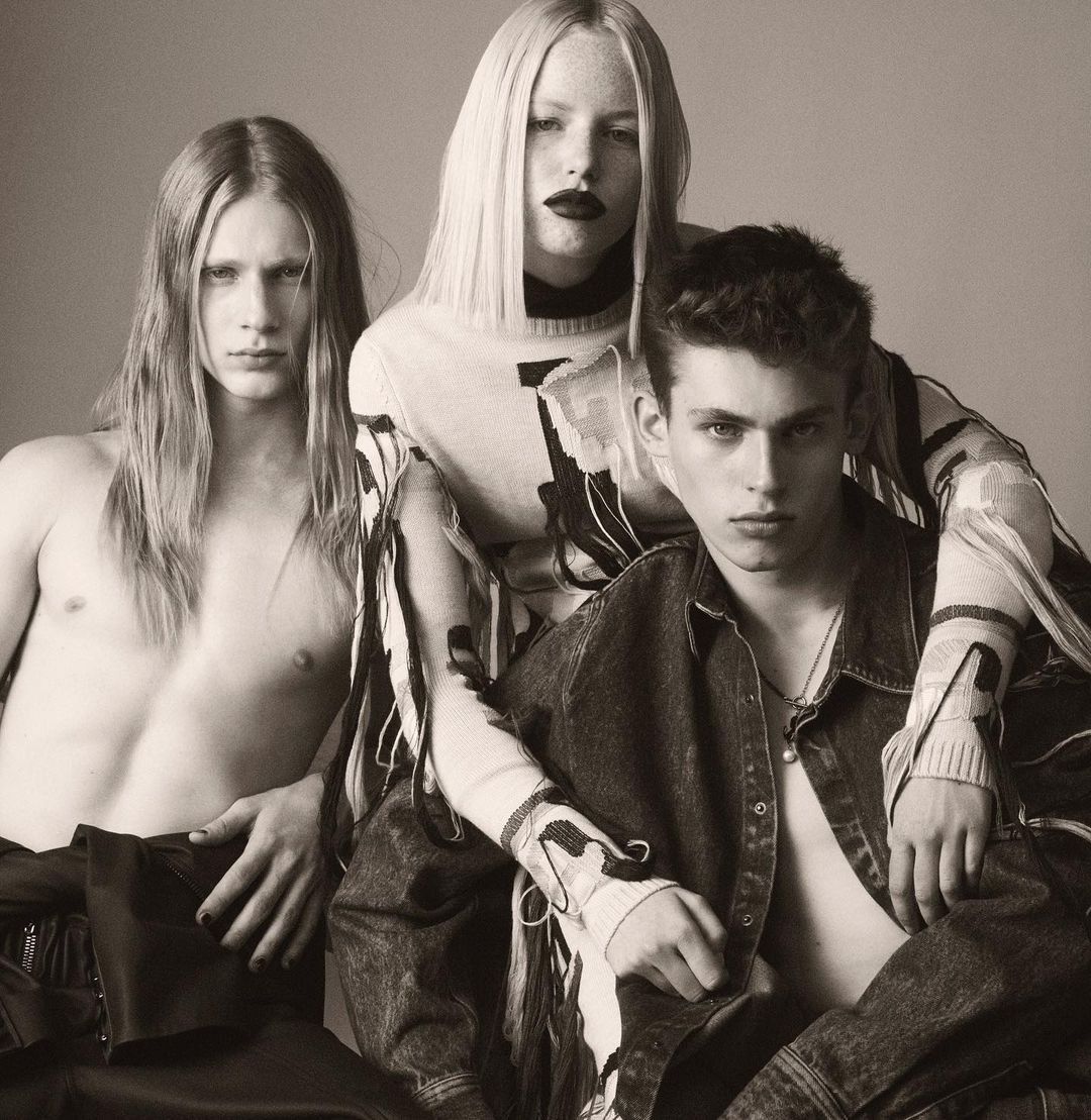 Three fashion models in a studio setting. A woman in the center with two men on either side, with neutral background and soft lighting.