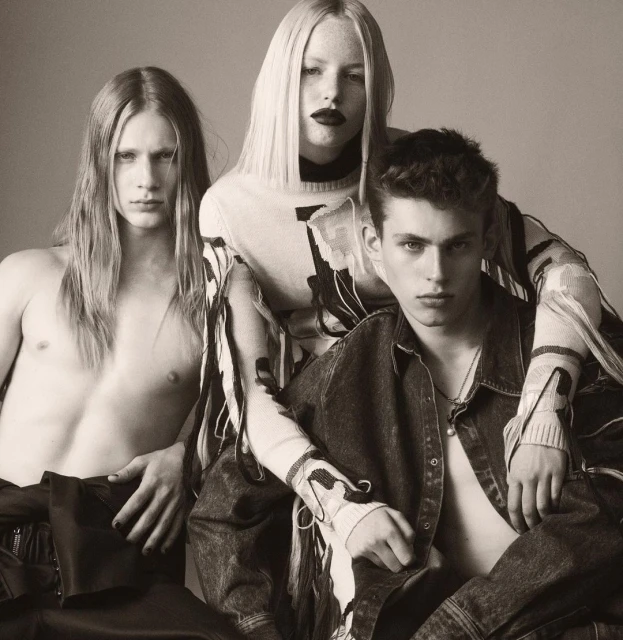 Three fashion models in a studio setting. A woman in the center with two men on either side, with neutral background and soft lighting.