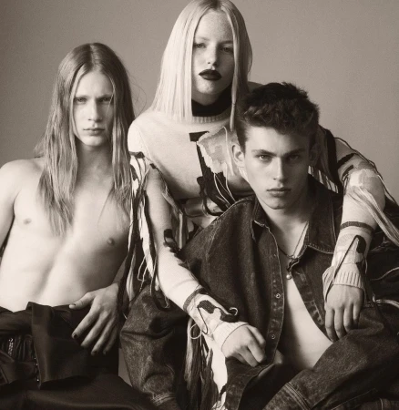 Three fashion models in a studio setting. A woman in the center with two men on either side, with neutral background and soft lighting.