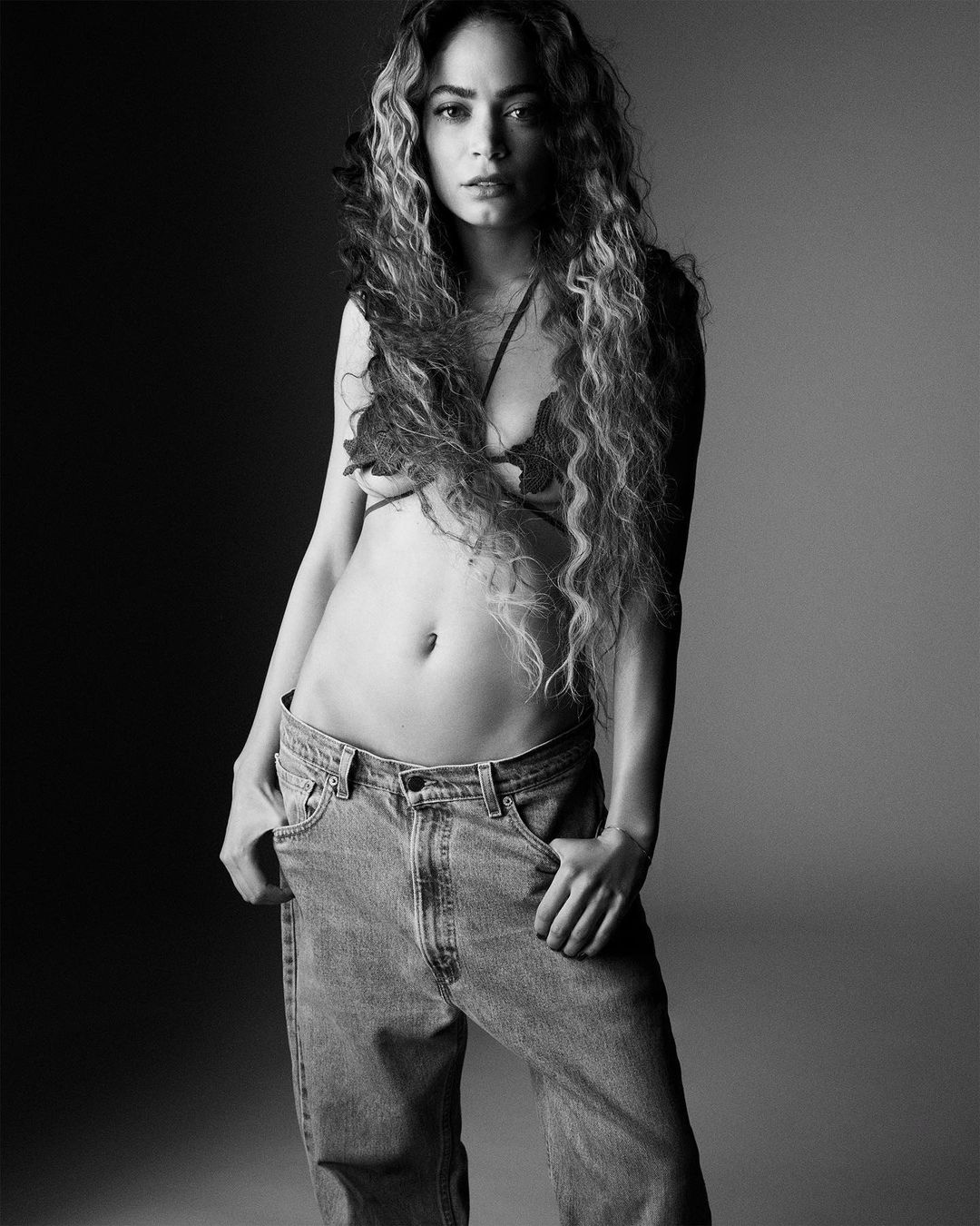 Monochrome portrait of a woman with long curly hair, wearing a leaf-patterned bra and baggy jeans, set against a graduated background.