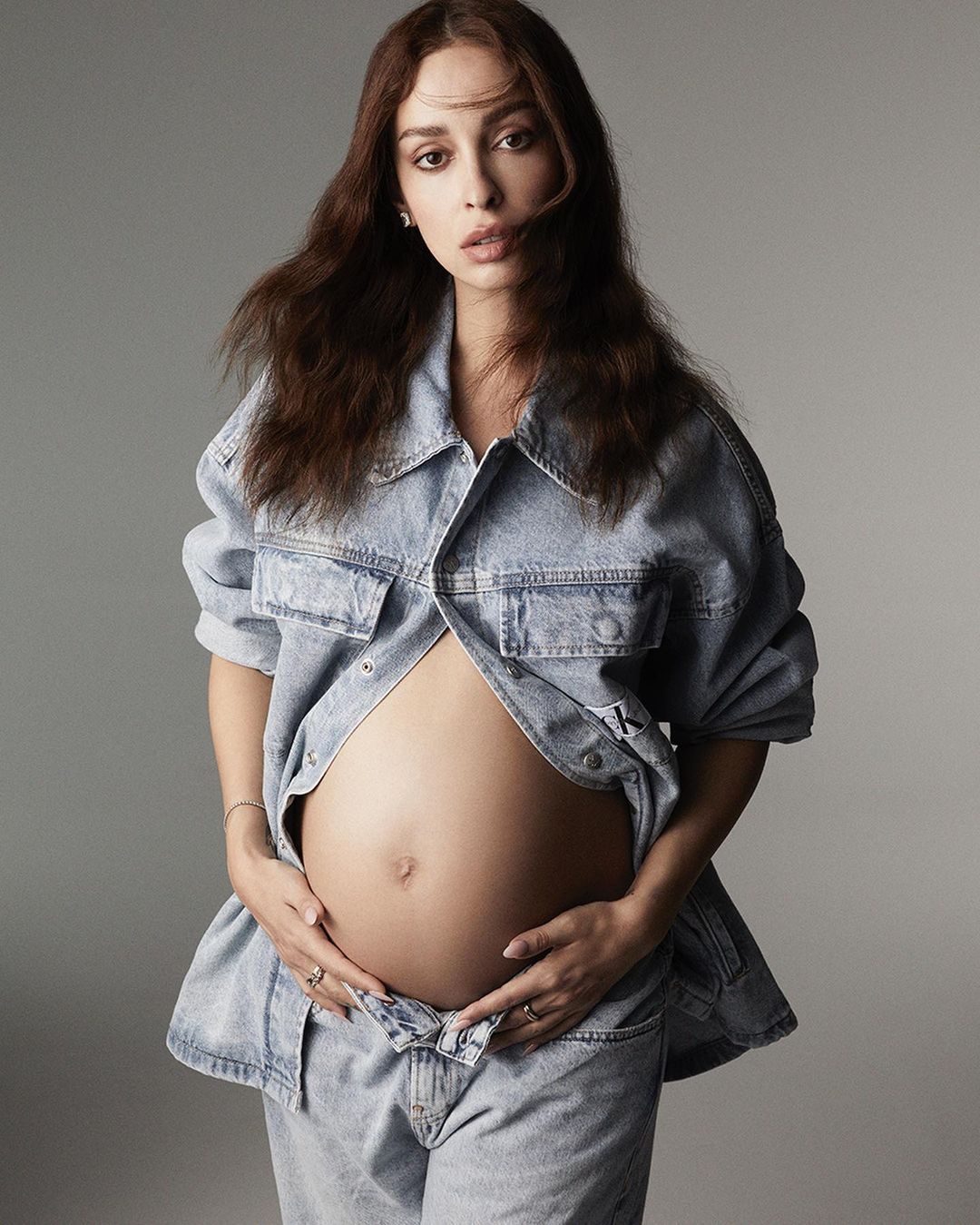 Pregnant woman with auburn hair wearing a denim jacket and jeans in a portrait against a grey background in a photo studio.