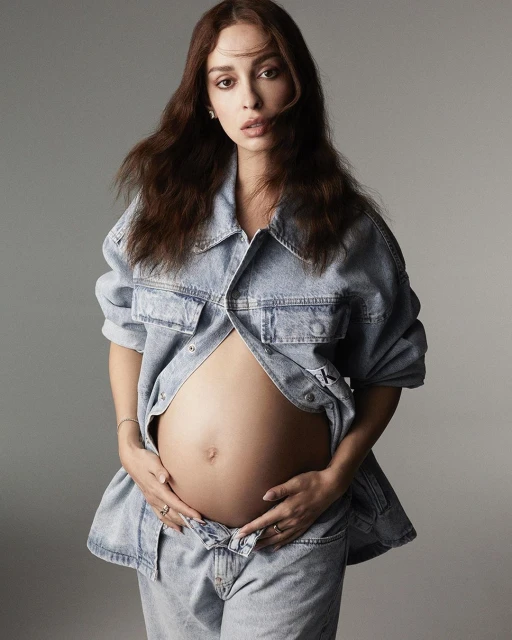 Pregnant woman with auburn hair wearing a denim jacket and jeans in a portrait against a grey background in a photo studio.