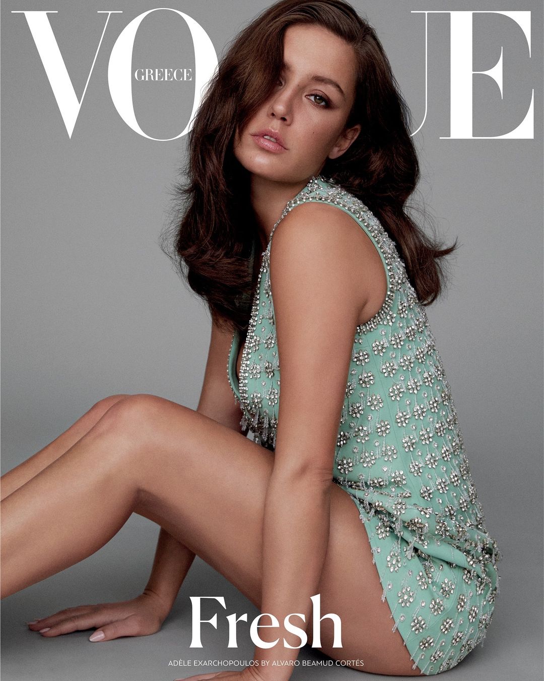 Brunette woman in embellished dress sits in vogue cover shot. Model gazes at viewer. Magazine cover features bold text. Fashion photography.