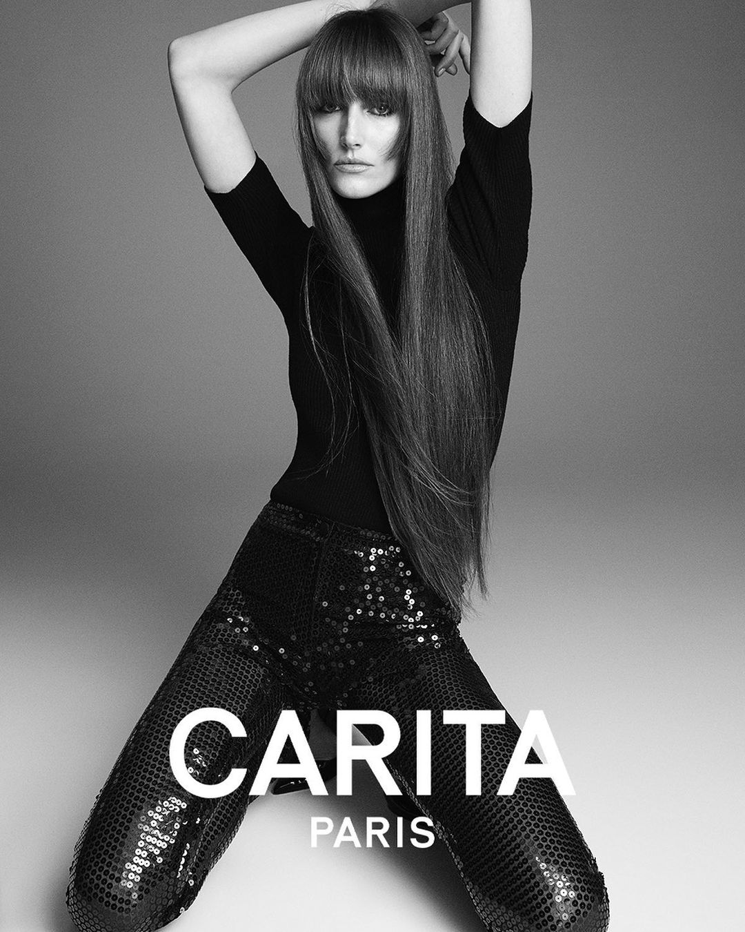Black and white fashion shot of a female model with long hair wearing a black turtleneck and sequined pants in a studio setting.